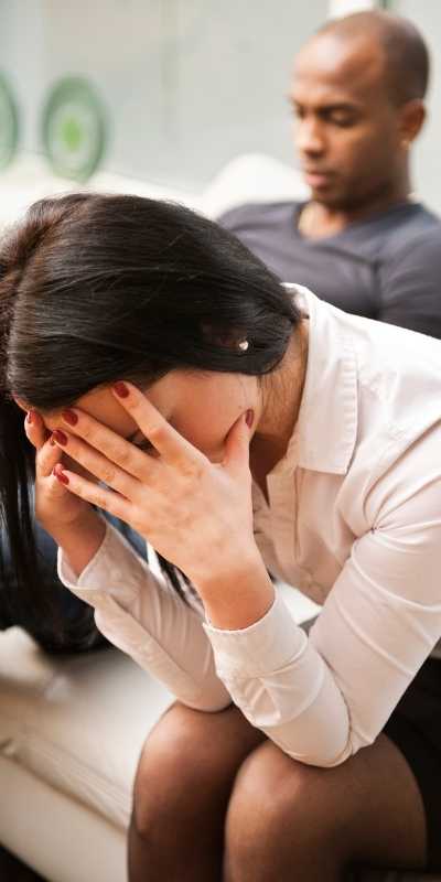 Woman with head in hands hunched over while her partner leans back against the couch behind her after a fight giving her the silent treatment in an unhealthy relationship